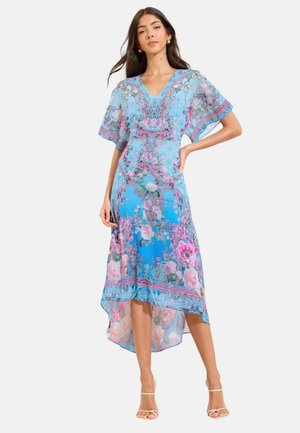 Vrouw draagt een blauw bloemen high-low jurk met korte mouwen en V-hals, staat met één hand op de heup en draagt witte sandalen met bandjes en hakken.