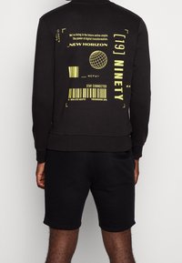 Svart sweatshirt med gul grafisk tryck, med text, jordklotdesign och streckkod. Matchas med svarta shorts, båda i mjukt material.