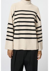 Béžový rolákový svetr s černými vodorovnými pruhy, žebrovanou texturou, širokými rukávy a volným střihem. Kombinován s černými kalhotami.