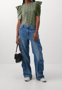 Top corto de manga corta verde texturizado con hombros abullonados, combinado con unos pantalones cargo azules sueltos. Un bolso negro y unas zapatillas blancas completan el look.