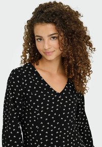 Jeune femme aux cheveux bouclés portant un haut noir à décolleté en V avec un petit motif de nœuds blancs, souriant doucement devant un fond uni.