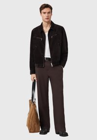 Καφέ μπουφάν από κορδόνι με δύο τσέπες στο στήθος με φερμουάρ, συνδυασμένο με λευκό t-shirt και καφέ παντελόνι. Κρατά μια τσάντα ώμου από ταμπά σουέντ.