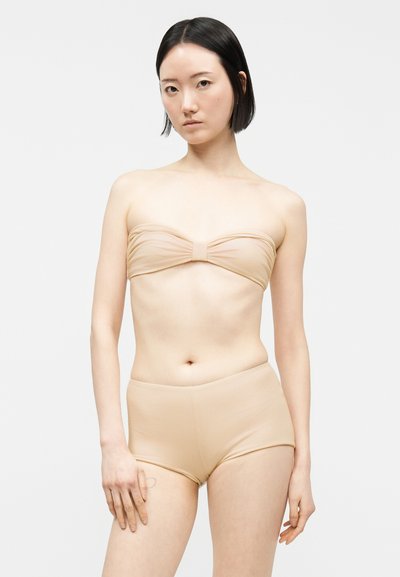Jeune femme aux cheveux noirs courts portant un haut bandeau beige et un short taille haute assorti sur un fond clair uni.