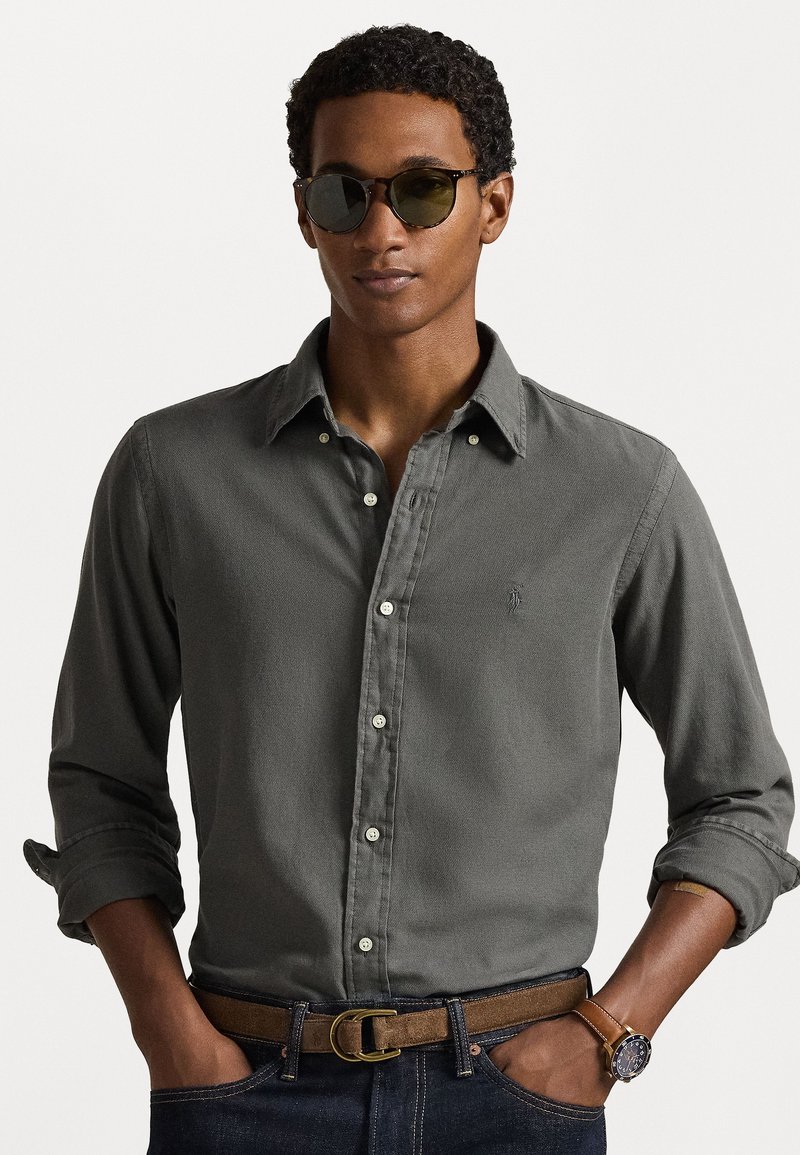 Tumši pelēks krekls ar piedurknēm, kas sarullētas, gaiši pogu akcenti, valkāts ar brūnu jostas. Modelis papildināts ar zaļām saulessargiem un pulksteni.