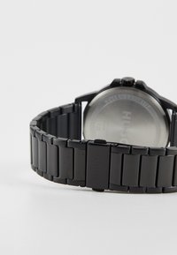 Zwart metalen horloge met een schakelarmbandontwerp, met een ronde wijzerplaat en een zilveren achterkant. Merken zichtbaar op de sluiting.