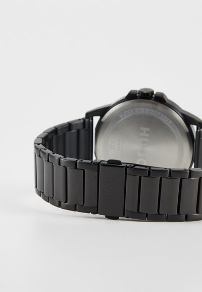Montre en métal noir avec un bracelet à maillons, présentant un cadran circulaire et un dos argenté. Branding visible sur le fermoir.