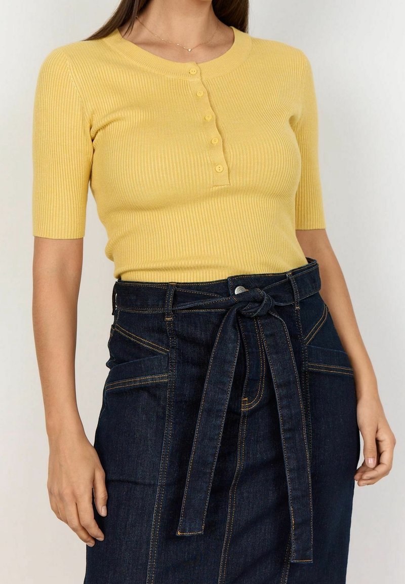 Top giallo a coste con maniche corte e dettagli con bottoni, abbinato a una gonna di denim scuro a vita alta con cintura annodata e tasche.