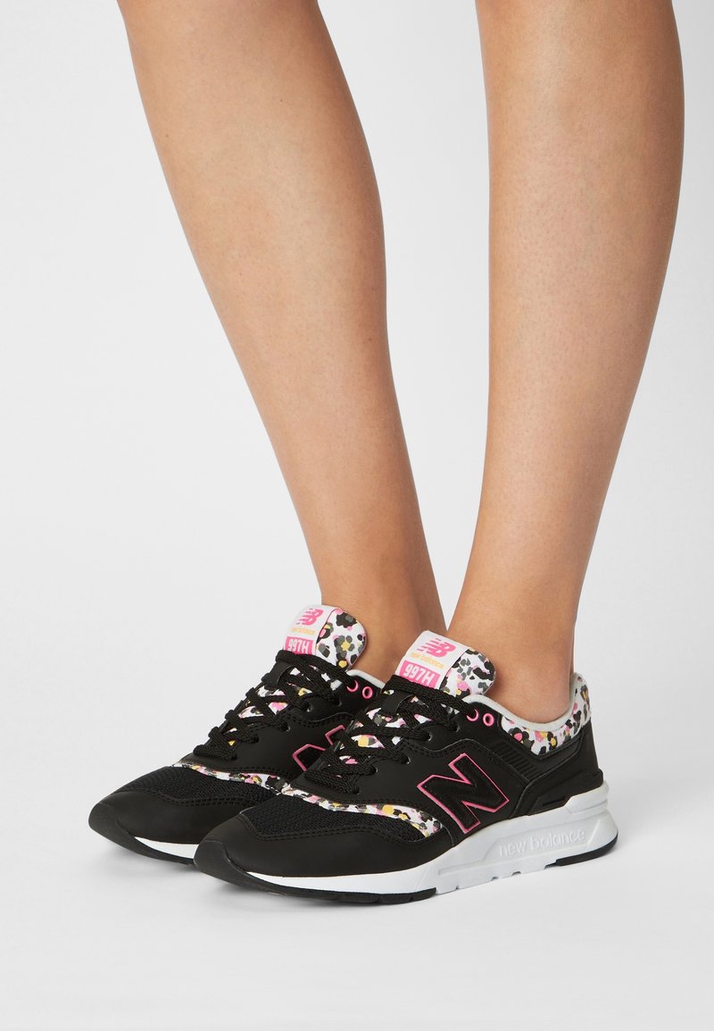 Jambes portant des baskets noires New Balance avec des accents roses et un motif léopard multicolore sur le col et la languette.