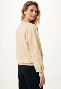 Sudadera beige claro de corte relajado, con puños acanalados y un pequeño logo en la parte posterior. Combinada con pantalones negros ajustados.