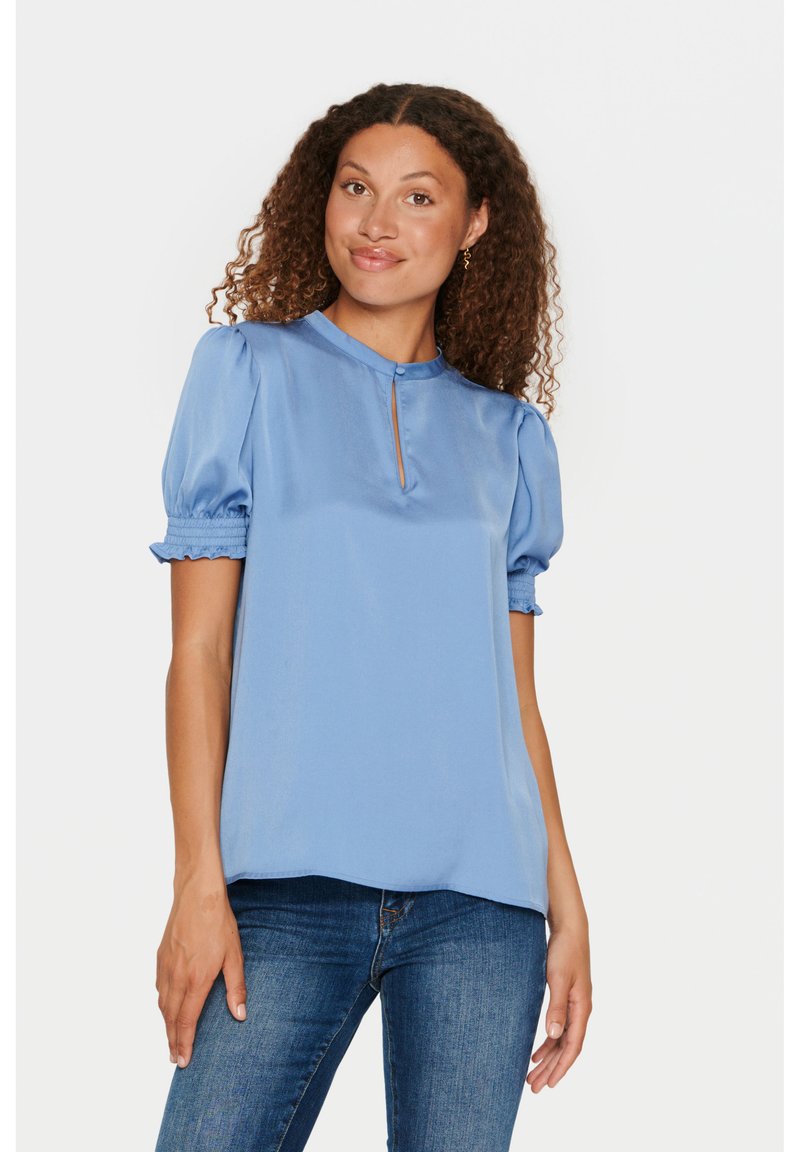 Blusa azzurra chiara con maniche a palloncino corte e scollatura a forma di chiave. Texture liscia, simile alla seta. Abbinata a jeans aderenti.