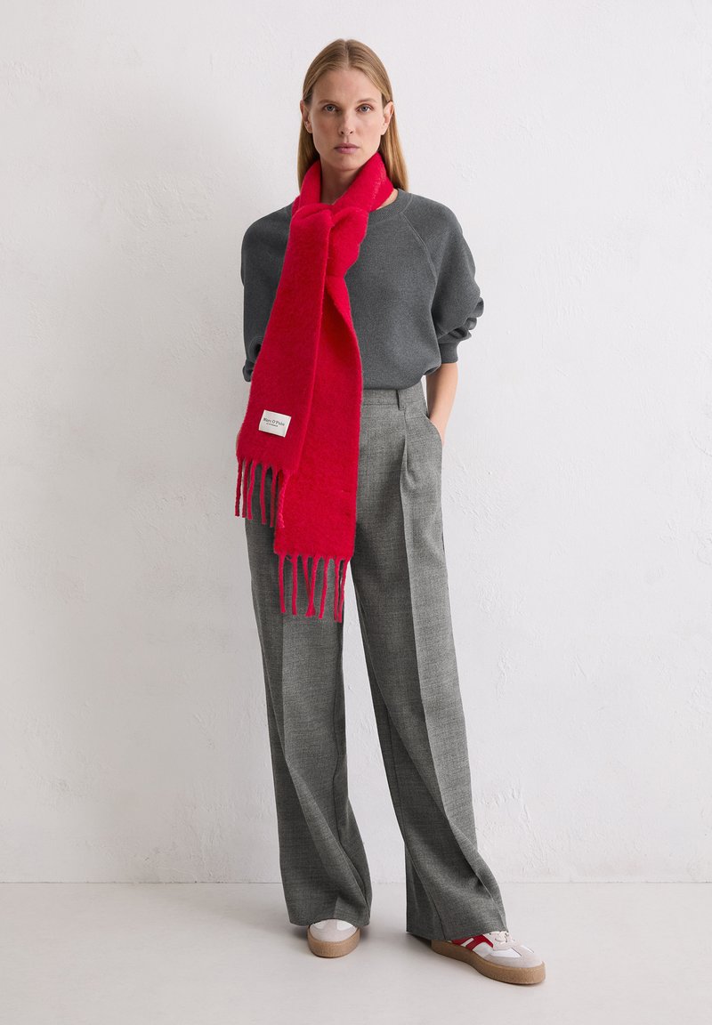 Roter Wollschal mit Fransen, kombiniert mit einem grauen Sweatshirt und grauen karierten Hosen mit lockerem Schnitt. Das Modell trägt Plateau-Schuhe.