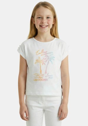 Fille souriante aux longs cheveux blonds, portant un t-shirt blanc avec un motif pastel de palmier et un pantalon blanc, debout devant un fond uni.