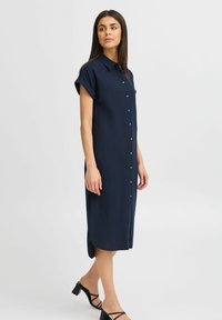 Rochie bleumarin cu nasturi, cu mâneci scurte, până la genunchi, realizată dintr-un material neted, având un tiv ușor curbat și un guler.