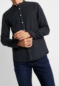 Camicia nera con bottoni bianchi a pois, con colletto a bandana, maniche lunghe e orlo curvo, abbinata a jeans blu scuro.