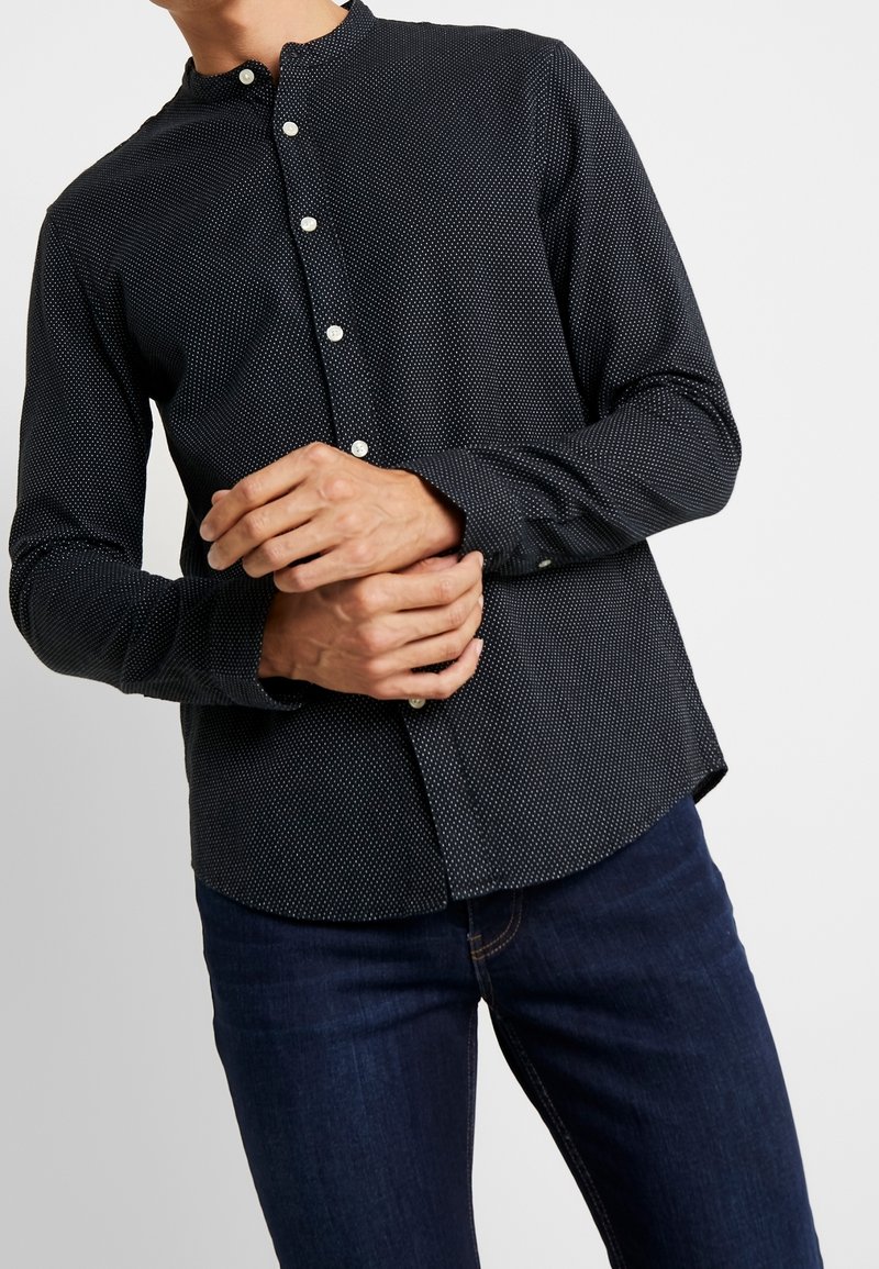 Camicia nera con bottoni bianchi a pois, con colletto a bandana, maniche lunghe e orlo curvo, abbinata a jeans blu scuro.