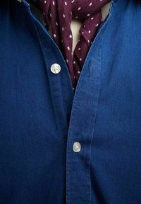 Tummansininen napitettavissa oleva paita, jossa on kaksi valkoista nappia ja viininpunainen huivi, jossa on valkoisia pisteitä, roikkumassa kaula-aukolla.