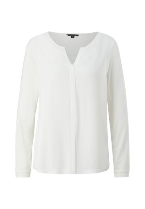 Kvinde iført en hvid langsleeve V-hals bluse, der er stoppet ned i beige bukser med brede ben og beige spidse sko, stående op ad en ensfarvet væg.