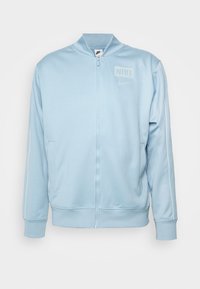 Giacca azzurra con zip in tessuto liscio, presenta un collo alto, strisce laterali bianche e un piccolo logo Nike sul petto.