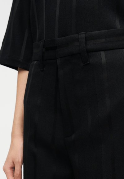 Juodos kelnės su subtiliomis horizontalios juostelėmis, su diržo kilpomis ir klostėmis, suteikiančiomis pritaikytą prigludimą, pagamintos iš tekstūruotos medžiagos.