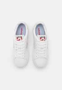 Paire de baskets blanches Levi's avec lacets, accents latéraux beige clair et logo rouge Levi's sur les semelles intérieures et les languettes.