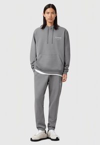 Grijze hoodie met trekkoorden en een kangoeroezak, gecombineerd met bijpassende grijze sweatpants. Beide hebben subtiele branding. Witte ondershirt zichtbaar.