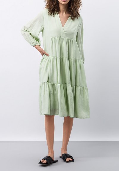 Robe tiered vert clair avec des manches longues, un décolleté en V et des poches latérales. Le matériau est doux avec une texture fluide. Portée avec des sandales noires.