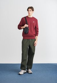 Bordo sweatshirt, zelene cargo hlače in sivi športni copati. Črna mini torbica čez ramo z naramnico. Enostaven dizajn, minimalni poudarki, sproščen kroj.