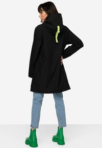 Manteau de pluie noir avec capuche, présentant un détail de sangle vert fluo. Associé à un jean bleu clair et à des bottes à talons chunky vertes.