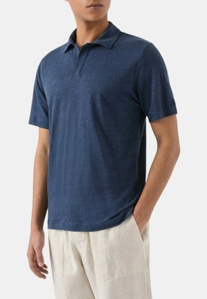 Uomo che indossa una polo a maniche corte blu scuro e pantaloni beige chiaro con una mano in tasca su uno sfondo semplice.