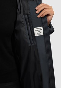 Zwarte gewatteerde jas met een gladde textuur. Het interieur toont een label met een slogan en website. Voorzien van een zijzak en ribbeldetails.