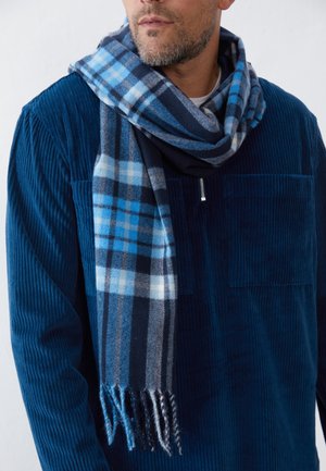 Mand iført en marineblå kordfløjlsjakke og blå ternet halstørklæde viklet omkring halsen mod en hvid baggrund.