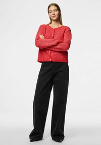 Röd stickad kappa med långa ärmar, rund halsringning och framknappar. Kombinerad med vida svarta byxor och spetsiga svarta skor.