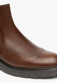 Bruine leren enkelboot met brogue-details, een gladde textuur, zij-elastische panelen en een gestructureerde rubberen zool.