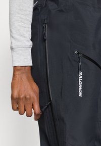 Calças pretas com um bolso lateral com fecho e textura lisa. Uma camisa de mangas longas cinza é visível. Apresenta um logo visível e fechos metálicos.