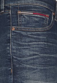 Tmavě modré džíny z denimu s texturovaným povrchem. Mají malou kapesní kapsu zdobenou červenými, b ílými a modrými pruhy. Je vidět detail měděného knoflíku.