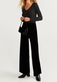 Pantalon noir à jambes larges en tissu texturé, associé à un top noir ajusté à col en V scintillant et un petit sac à main noir. Bottines brillantes.