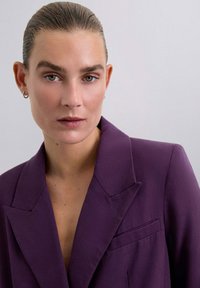 Blazer ajustado morado con solapa marcada, bolsillo en el pecho y hombros estructurados. Tela suave con detalles de textura mínimos.
