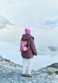 Puchowy fioletowy płaszcz, białe spodnie i brązowe buty; różowa dzianinowa czapka, kolorowy plecak z różowymi, niebieskimi i żółtymi akcentami na śnieżnym tle.