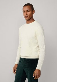 Suéter de punto cableado color crema con cuello redondo, dobladillo y puños acanalados, combinado con pantalones de pana de color verde oscuro. Diseño simple y texturizado.