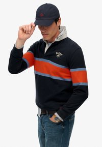 Marineblauwe lange mouwen polo met oranje en blauwe horizontale strepen, met een kraagontwerp en geborduurd logo. Draag het met spijkerbroeken en een pet.