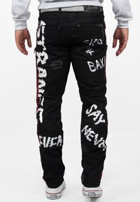 Jeans in denim nero con grafiche testuali bianche. Dettagli strappati e accenti laterali rossi. Indossati con sneaker bianche.