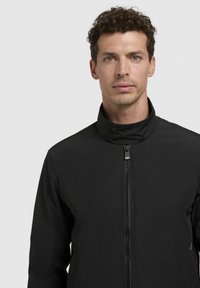 Black jacket featuring a high collar, zip closure, and snap buttons. The material appears smooth with a minimalistic design and no visible patterns.
