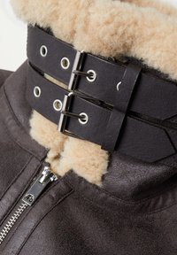 Gros plan sur le col d'une veste en cuir marron foncé avec doublure en peau lainée beige, fermeture éclair argentée et deux sangles en cuir noir avec boucles en métal.