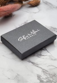 Graue rechteckige Schmuckbox mit strukturierten Oberflächen und silbernem Logo auf der Oberseite. Auf einem hellen Marmorhintergrund platziert.