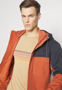 Giacca arancione e blu navy con cappuccio regolabile, dotata di chiusura con zip e tasche. La camicia a strati sottostante presenta strisce orizzontali di vari colori.