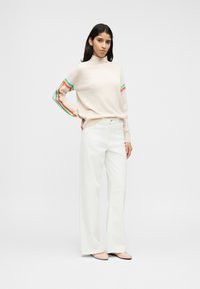 Πλεκτό πουλόβερ σε ανοιχτό μπεζ με πολύχρωμες ρίγες στα μανίκια, συνδυασμένο με λευκά παντελόνια φαρδιάς γραμμής και μπεζ φλατ παπούτσια.