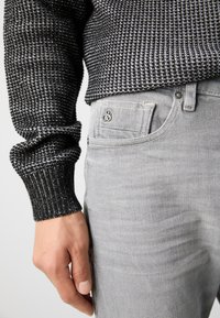 Persoană îmbrăcată în blugi gri deschis și un pulover cu un model alb-negru, cu mâna relaxată lângă corp, concentrându-se pe zona buzunarului pantalonilor.
