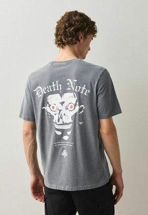 Ung mand med krøllet hår iført grå "Death Note" T-shirt med hvid kranium med røde øjne og tekst på ryggen, stående mod en ensfarvet baggrund.