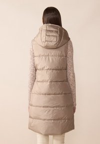 Beige puffer-väst med en stor huood, som har horisontella quiltade sektioner och en längre bakre fåll. Lätt tyg med en slät textur.