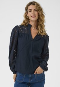 Blouse bleu marine avec manches longues en dentelle, col haut et texture subtile. Corsage structuré avec un détail froncé et des poignets ajustés.